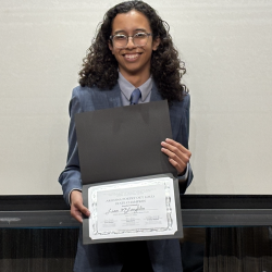 A young person in a suit with long curly hair holds a certificate